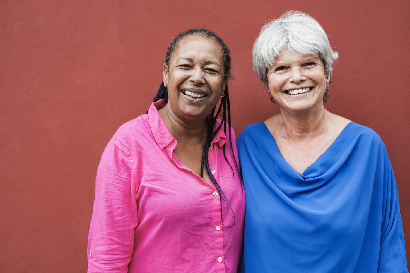 Two older women smiling in front of a red wall, enjoy the benefits of photobiomodulation in Littleton.